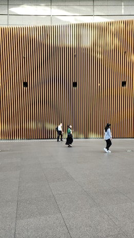 A modern architectural wall featuring vertical wooden slats with several circular speakers or installations evenly spaced. Three people are walking across a smooth, spacious floor in front of the wall, each dressed casually.