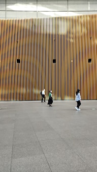 A modern architectural wall featuring vertical wooden slats with several circular speakers or installations evenly spaced. Three people are walking across a smooth, spacious floor in front of the wall, each dressed casually.