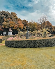 A well-maintained garden features carefully trimmed hedges and vibrant flowering plants. Statues are situated amidst the greenery, and tall trees in the background display autumn foliage. The sky is partly cloudy, adding contrast to the scene.