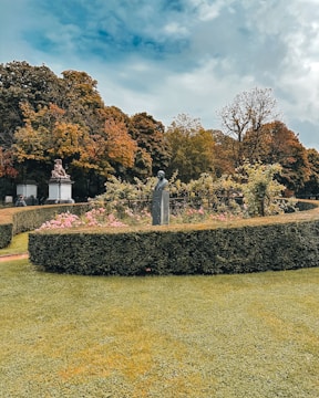 A well-maintained garden features carefully trimmed hedges and vibrant flowering plants. Statues are situated amidst the greenery, and tall trees in the background display autumn foliage. The sky is partly cloudy, adding contrast to the scene.