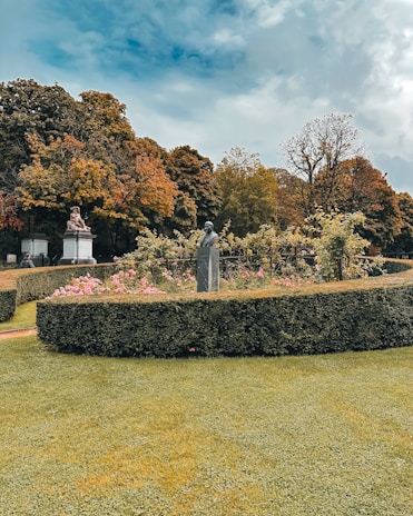 A well-maintained garden features carefully trimmed hedges and vibrant flowering plants. Statues are situated amidst the greenery, and tall trees in the background display autumn foliage. The sky is partly cloudy, adding contrast to the scene.