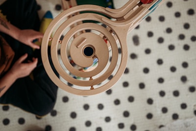 A wooden spiral toy track is positioned over a dotted background. A person sits cross-legged nearby, partially visible, focusing on the toy.