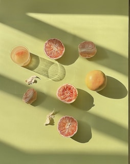 Sliced grapefruit and a whole grapefruit are arranged on a light green surface. Soft shadows create a pattern across the scene, and a glass of juice is positioned on the left, casting its own shadow.