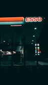 A dimly lit gas station at night featuring a prominent ESSO sign. The area is mostly dark, with some illuminated sections highlighting gas pumps and a store in the background. Vertical bars in the foreground add a sense of separation or framing.