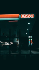 A dimly lit gas station at night featuring a prominent ESSO sign. The area is mostly dark, with some illuminated sections highlighting gas pumps and a store in the background. Vertical bars in the foreground add a sense of separation or framing.