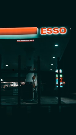 A dimly lit gas station at night featuring a prominent ESSO sign. The area is mostly dark, with some illuminated sections highlighting gas pumps and a store in the background. Vertical bars in the foreground add a sense of separation or framing.