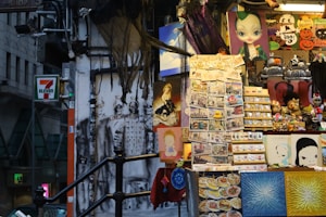A vibrant market stall features a variety of items including colorful postcards, artistic prints, and small souvenirs. Halloween-themed decorations can be seen, including black and orange signage with skull imagery. There is artwork depicting animated or abstract faces, alongside a display of cartoonish figures. A 7-Eleven sign is visible in the background amidst an urban setting with buildings.
