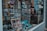 A storefront window displays an assortment of postcards and promotional materials. The interior is filled with racks of neatly arranged greeting cards, with various designs labeled 'Happy Birthday' among others. Packs of promotional Lyca Mobile SIM cards and brochures are visible on the window ledge. Reflections of nearby buildings and pedestrians are visible on the window, adding depth to the scene.
