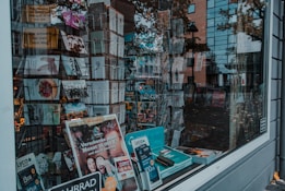 A storefront window displays an assortment of postcards and promotional materials. The interior is filled with racks of neatly arranged greeting cards, with various designs labeled 'Happy Birthday' among others. Packs of promotional Lyca Mobile SIM cards and brochures are visible on the window ledge. Reflections of nearby buildings and pedestrians are visible on the window, adding depth to the scene.