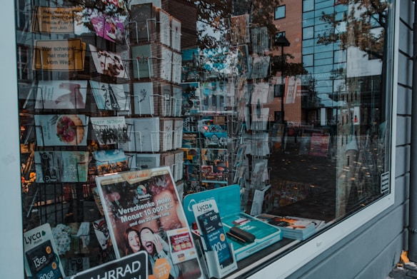 A storefront window displays an assortment of postcards and promotional materials. The interior is filled with racks of neatly arranged greeting cards, with various designs labeled 'Happy Birthday' among others. Packs of promotional Lyca Mobile SIM cards and brochures are visible on the window ledge. Reflections of nearby buildings and pedestrians are visible on the window, adding depth to the scene.