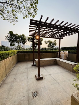 A patio area with a wooden pergola structure casting shadows on the tiled floor. Surrounding the space are green leafy plants, with the sun shining through the foliage. A stack of white plastic chairs is situated in the corner.