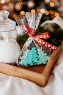 A wooden tray holds items for a festive setting. Inside, there's a clear plastic bag tied with a red ribbon, containing turquoise Christmas tree-shaped candies or confections. In the background, a white ceramic jar with a lid is visible, as well as blurred out warm glowing lights and hints of Christmas tree foliage, suggesting a cozy holiday atmosphere.