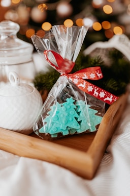 A wooden tray holds items for a festive setting. Inside, there's a clear plastic bag tied with a red ribbon, containing turquoise Christmas tree-shaped candies or confections. In the background, a white ceramic jar with a lid is visible, as well as blurred out warm glowing lights and hints of Christmas tree foliage, suggesting a cozy holiday atmosphere.