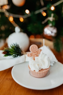 A festive cupcake topped with white frosting and a gingerbread man candy sits on a white plate. In the background, a decorated Christmas tree with blurred lights and a wrapped gift or napkin is visible, creating a warm holiday atmosphere.