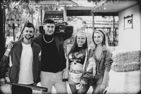 A group of four individuals is posing together. One person is holding a large boombox on their shoulder. Another person is carrying a baseball bat. The group appears to be dressed in a retro style with headbands and casual outfits.