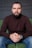 A man with a beard sitting on a green couch, wearing a brown turtleneck sweater and blue jeans. His hands are clasped, and he is wearing a large wristwatch. The background is a gray wall.