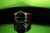 A wristwatch with a black dial and white numerals is prominently displayed. It features a brown strap and is positioned against a reflective surface, creating a mirror-like effect beneath it. The background is a gradient of bright green transitioning to dark, adding a striking contrast to the watch.