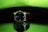 A wristwatch with a black dial and white numerals is prominently displayed. It features a brown strap and is positioned against a reflective surface, creating a mirror-like effect beneath it. The background is a gradient of bright green transitioning to dark, adding a striking contrast to the watch.