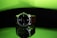 A wristwatch with a black dial and white numerals is prominently displayed. It features a brown strap and is positioned against a reflective surface, creating a mirror-like effect beneath it. The background is a gradient of bright green transitioning to dark, adding a striking contrast to the watch.
