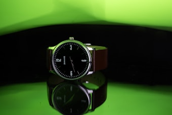 A wristwatch with a black dial and white numerals is prominently displayed. It features a brown strap and is positioned against a reflective surface, creating a mirror-like effect beneath it. The background is a gradient of bright green transitioning to dark, adding a striking contrast to the watch.