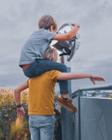 Two children are engaged in a playful activity at an observation point. One child is wearing a gray shirt and dark pants, perched on the shoulders of the other child, who is wearing a mustard yellow shirt and light blue jeans. They are interacting with a large outdoor telescope. In the background, there are ornamental grasses and a cloudy sky, suggesting an outdoor setting.