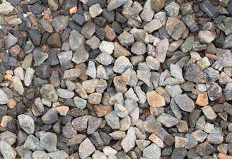 A large collection of irregularly shaped rocks and stones in various shades of gray, brown, and beige is closely packed together. The stones vary in size and texture, creating a natural and rugged surface.
