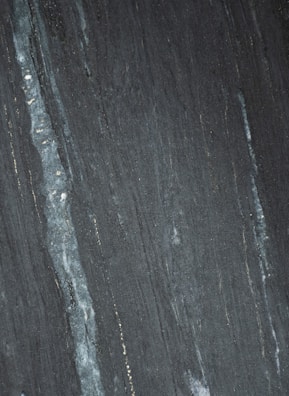 Dark stone surface with vertical striations in shades of gray and black. Subtle hints of lighter streaks and fine textures create a natural pattern.