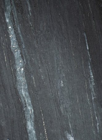 Dark stone surface with vertical striations in shades of gray and black. Subtle hints of lighter streaks and fine textures create a natural pattern.