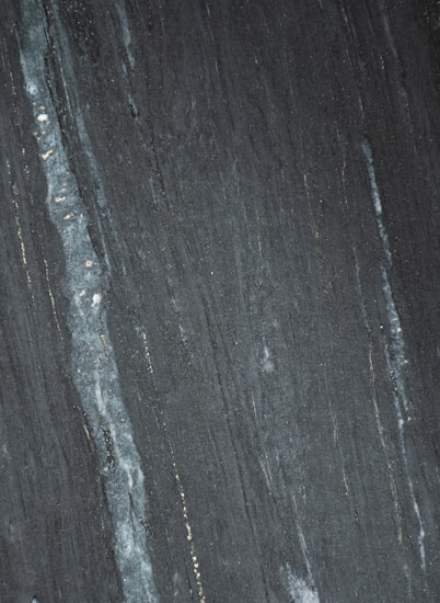 Dark stone surface with vertical striations in shades of gray and black. Subtle hints of lighter streaks and fine textures create a natural pattern.