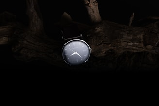 A sleek wristwatch is illuminated against a dark, rustic background featuring textured wood. The watch face is clear with minimalistic markings and hands, creating a contrast with the shadowy environment.