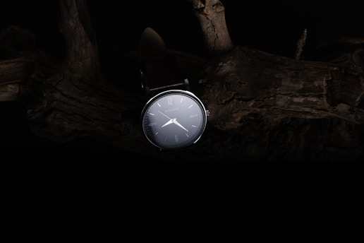 A sleek wristwatch is illuminated against a dark, rustic background featuring textured wood. The watch face is clear with minimalistic markings and hands, creating a contrast with the shadowy environment.