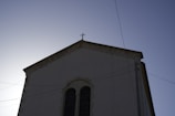 A building with a cross on the roof, partially silhouetted against a clear sky. The facade is simple with large windows and electrical wires crossing in front.