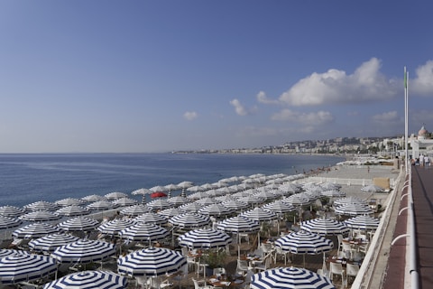 Numerous blue and white striped beach umbrellas are neatly arranged on a pebbled shoreline next to a calm sea under a clear blue sky. In the background, a city with numerous buildings stretches along the coastline. A vibrant red umbrella stands out among the others.