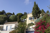 A picturesque scene of a Mediterranean-style house surrounded by lush greenery and vibrant flowers. The house features stucco walls and blue shutters, blending harmoniously with the surrounding cypress and palm trees. Bright bougainvillea adds a splash of color near the entrance.