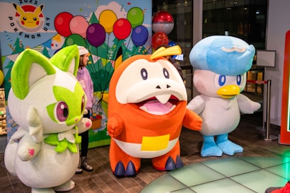 Large plush character costumes of various Pokémon are displayed indoors with a person standing nearby. The setting includes colorful balloons and a festive Pokémon-themed backdrop.