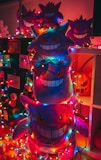 Plush toys shaped like a ghost character are stacked on top of each other. They are surrounded by colorful string lights, creating a festive and whimsical atmosphere. The toys have a mischievous and playful grin, and one can see a carved pumpkin in the background.