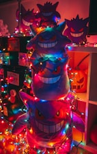 Plush toys shaped like a ghost character are stacked on top of each other. They are surrounded by colorful string lights, creating a festive and whimsical atmosphere. The toys have a mischievous and playful grin, and one can see a carved pumpkin in the background.