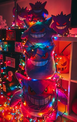 Plush toys shaped like a ghost character are stacked on top of each other. They are surrounded by colorful string lights, creating a festive and whimsical atmosphere. The toys have a mischievous and playful grin, and one can see a carved pumpkin in the background.