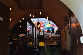 A cozy, dimly lit hallway leads into a vibrant outdoor space filled with colorful umbrellas hanging overhead. Warm, vintage-style light bulbs dangle from the ceiling. There is a sense of warmth and invitation with an 'open' sign visible, and various decorations and plants add to the atmosphere.