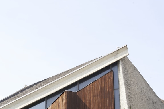 A modern architectural structure with an angular roofline combining concrete and wooden elements. The facade features a textured, gray concrete surface with a prominent wooden paneling section, contrasted by a clear blue sky in the background.