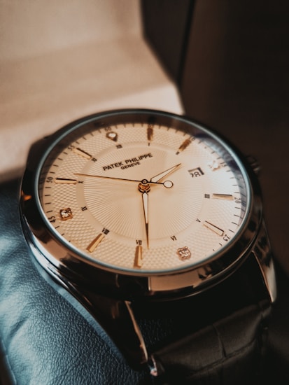 A luxury wristwatch with a sleek, polished metal case and a detailed, textured dial. The watch has gold hour markers, including Roman numerals and diamond accents at select positions. The hands are finely crafted and the brand name is visible on the dial.