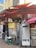 The image depicts a street food stall with colorful awnings in a busy alley. There are signs with Korean text and various images, including food menus and promotional materials. A glass refrigerator displays various packaged food items, and there are crates and cooking utensils nearby.