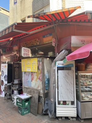 The image depicts a street food stall with colorful awnings in a busy alley. There are signs with Korean text and various images, including food menus and promotional materials. A glass refrigerator displays various packaged food items, and there are crates and cooking utensils nearby.