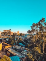 An urban landscape featuring a mix of densely packed residential buildings with vibrant rooftop colors. Tall trees are scattered throughout, providing a natural contrast to the city environment. In the background, large buildings rise against a clear blue sky.