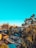 An urban landscape featuring a mix of densely packed residential buildings with vibrant rooftop colors. Tall trees are scattered throughout, providing a natural contrast to the city environment. In the background, large buildings rise against a clear blue sky.
