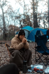 A person is sitting wrapped in a blanket in a forest setting, appearing relaxed and contemplative. They are wearing a beanie and are surrounded by camping gear, such as a blue tent and folding chairs. The ground is covered with dry leaves, indicating an autumn setting.