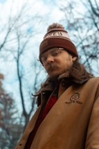A person wearing a brown jacket with a logo and text 'Skeet & Trap', along with a red and white knit hat. The individual appears to have long hair and glasses, standing in an outdoor setting with leafless trees and a cloudy sky in the background.