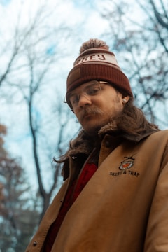A person wearing a brown jacket with a logo and text 'Skeet & Trap', along with a red and white knit hat. The individual appears to have long hair and glasses, standing in an outdoor setting with leafless trees and a cloudy sky in the background.