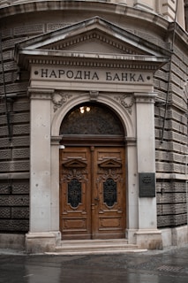 An ornate wooden double door set within a grand stone archway, featuring intricate carvings and metalwork. The building exterior has a classical architectural style with decorative stone elements and a sign above the door with Cyrillic text.