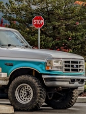 A teal and silver Ford F-250 truck is parked next to a stop sign. The truck appears rugged with large off-road tires and is positioned in a natural setting with dense green foliage and some red leaves in the background.